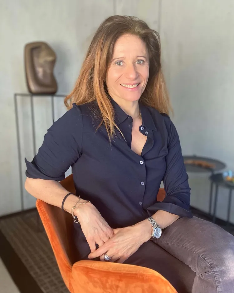 Portrait photographique d’Anne Zürcher Kondratowicz, hypnothérapeute à Fribourg, souriante, assise dans un fauteuil orange, en chemise bleu marine.