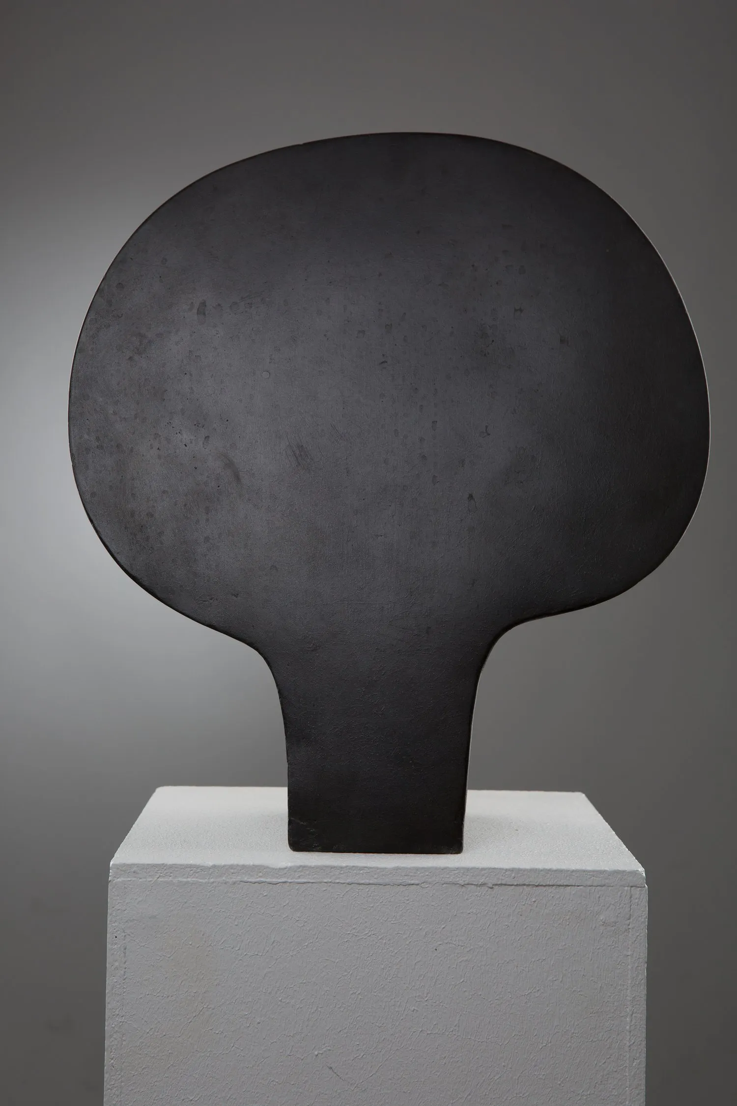 « Spiegel I » de Jan Hegy : sculpture en métal noir mat, large forme ronde et lisse portée par un pied étroit, posée sur un socle blanc.